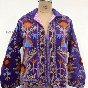 Peut inclure: Veste en velours violet avec broderies florales orange, bleu et rouge. La veste a un large col et des manches longues. La doublure est rayée rose et blanc. La veste est présentée sur un mannequin.