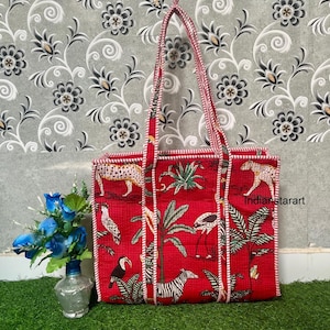 May include: A red tote bag with a white trim and handles. The bag features a repeating pattern of zebras, leopards, birds, and palm trees. The bag is sitting on a green surface with a floral arrangement.