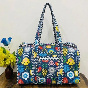 May include: A quilted duffel bag featuring a dark blue base with a multi-colored floral and leaf pattern in yellow, red, light blue, and white. The bag includes two long shoulder straps, a front zippered pocket, and a striped trim along the edges.