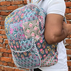 Puede incluir: Una mochila gris de patchwork con estampados florales y ribetes blancos. La mochila tiene un bolsillo grande en la parte delantera y correas ajustables.