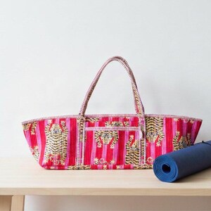 May include: A red and pink striped tote bag with tiger designs and two handles. A rolled-up blue yoga mat rests beside the bag on a wooden surface. The bag has a rectangular shape and a small pocket.