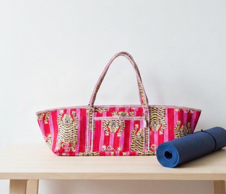 May include: A red and pink striped tote bag with tiger designs and two handles. A rolled-up blue yoga mat rests beside the bag on a wooden surface. The bag has a rectangular shape and a small pocket.