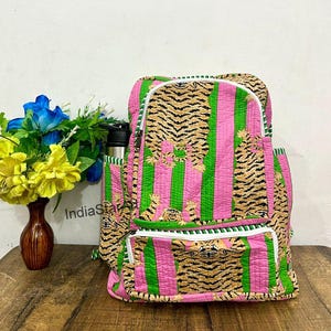 May include: A quilted backpack with a vibrant design. It features alternating pink and green stripes, accented with tiger print panels. The backpack has a white zipper and a side pocket for a water bottle. A vase of flowers sits nearby.