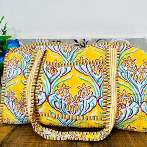 Puede incluir: Una bolsa de lona acolchada amarilla con estampado floral. La bolsa tiene un ribete a rayas marrón y blanco y dos asas.