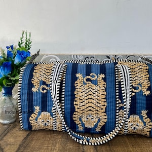 Puede incluir: Una bolsa de lona azul y beige con estampado de tigre y rayas verticales. La bolsa presenta un patrón repetido de tigres y está adornada con tela a rayas blancas y negras. La bolsa tiene dos asas y una correa para el hombro.