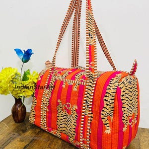 May include: A brightly coloured duffel bag with orange and pink stripes and tiger print details. The bag has long, striped handles and a rectangular shape. The bag is made of patterned fabric and is on a wooden surface.