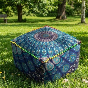 Puede incluir: Puf cuadrado azul con un diseño de mandala en tonos turquesa, turquesa y morado. Los bordes están adornados con pompones de colores. Este cojín de suelo decorativo es ideal para añadir un toque bohemio a cualquier espacio.