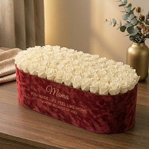 May include: A burgundy velvet floral arrangement box filled with ivory roses. The box has the word "Mama" and a message printed on the front. A gold vase with greenery is in the background.