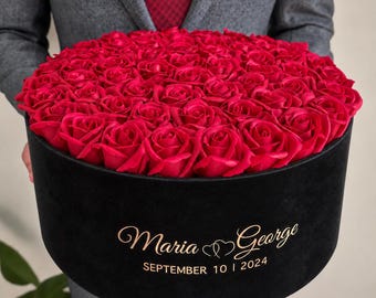 May include: A large round black velvet box filled with numerous vibrant red roses. The front of the box features gold lettering that reads Maria George and SEPTEMBER 10 | 2024, separated by a small heart icon between the two names.