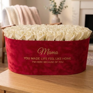 May include: A red velvet oval flower box filled with white roses. The box has gold text that reads "Mama" and a message below. The arrangement sits on a wooden surface.