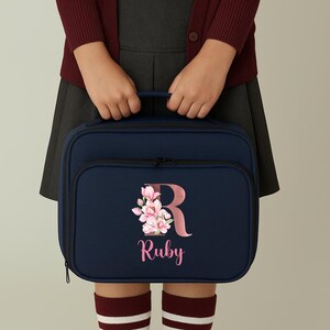 Puede incluir: Bolsa de almuerzo azul marino con un bolsillo frontal y una cremallera negra. La bolsa presenta un dise&ntilde;o floral con la letra "R" y el nombre "Ruby" en rosa. La bolsa es sostenida por una persona que lleva una rebeca granate y una falda gris.