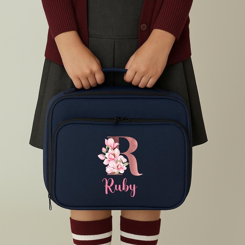 Puede incluir: Bolsa de almuerzo azul marino con un bolsillo frontal y una cremallera negra. La bolsa presenta un dise&ntilde;o floral con la letra "R" y el nombre "Ruby" en rosa. La bolsa es sostenida por una persona que lleva una rebeca granate y una falda gris.