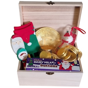Puede incluir: Una caja de madera llena de regalos con tem&aacute;tica navide&ntilde;a. La caja contiene un par de calcetines navide&ntilde;os verdes y rojos, una cuchara de madera de Pap&aacute; Noel, una cuchara de madera de elfo, una moneda de chocolate dorada, una barra de chocolate Cadbury Dairy Milk Buttons y una figura de mu&ntilde;eco de nieve.