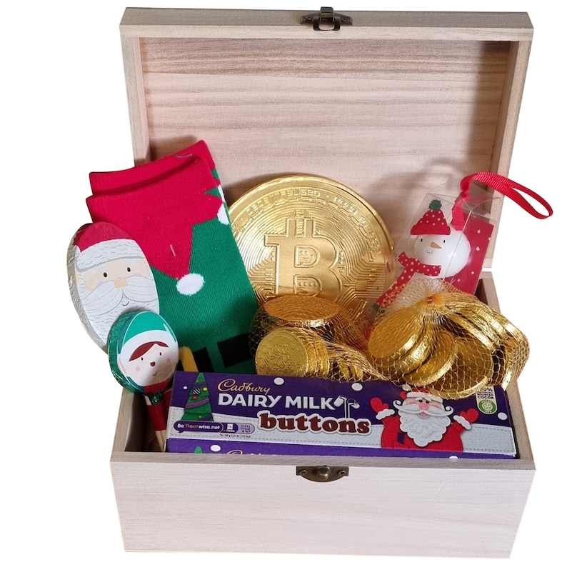 Puede incluir: Una caja de madera llena de regalos con tem&aacute;tica navide&ntilde;a. La caja contiene un par de calcetines navide&ntilde;os verdes y rojos, una cuchara de madera de Pap&aacute; Noel, una cuchara de madera de elfo, una moneda de chocolate dorada, una barra de chocolate Cadbury Dairy Milk Buttons y una figura de mu&ntilde;eco de nieve.