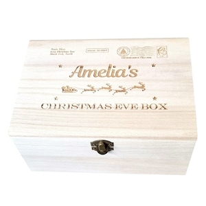 Puede incluir: Una caja de madera blanca con una tapa con bisagras. La tapa est&aacute; decorada con una inscripci&oacute;n personalizada que dice "Amelia's Christmas Eve Box". La inscripci&oacute;n est&aacute; rodeada de estrellas y una representaci&oacute;n del trineo de Pap&aacute; Noel con renos.