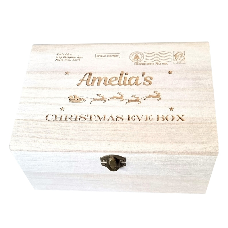 Puede incluir: Una caja de madera blanca con una tapa con bisagras. La tapa est&aacute; decorada con una inscripci&oacute;n personalizada que dice "Amelia's Christmas Eve Box". La inscripci&oacute;n est&aacute; rodeada de estrellas y una representaci&oacute;n del trineo de Pap&aacute; Noel con renos.