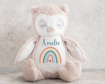 Op de afbeelding: Een zacht, beige en wit pluche uil-speelgoed. De uil heeft gesloten ogen en een regenboogontwerp met een hart op de borst, met de naam "Amelie" erboven. Een schattig en knuffelbaar kinderspeelgoed.