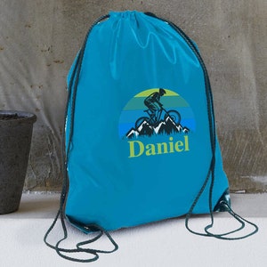 May include: A turquoise blue drawstring bag featuring a graphic of a person riding a mountain bike in front of a sunset and mountains. The text "Daniel" is printed below the graphic.
