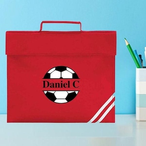May include: Red personalised book bag with a white football graphic and the name "Daniel C" printed in black.