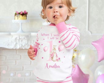 May include: A white and pink striped pajamas with a pink bunny rabbit illustration and the text "When I wake I will be 1 Amelia".