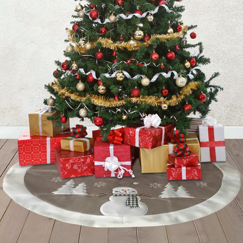 May include: A round, taupe-coloured Christmas tree skirt featuring a central white snowman appliqu&eacute; wearing a black and white chequered scarf, surrounded by white embroidered pine trees and snowflakes, finished with a wide, solid white border trim.