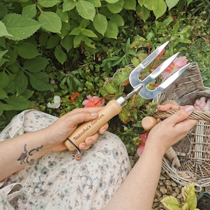 May include: A close-up of a gardening tool with a wooden handle. The handle is engraved with the words "Natasha's Garden". The tool has three tines and is made of metal.