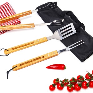 May include: A barbecue tool set with wooden handles and stainless steel heads, including a spatula, fork, and tongs. The handles are engraved with text. A red and white checkered cloth, a black carrying case, a red pepper, and cherry tomatoes are also present.