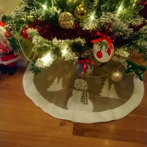 May include: A circular Christmas tree skirt featuring a light brown centre with a white snowman illustration wearing a plaid scarf, surrounded by white pine tree silhouettes and snowflakes, finished with a wide white border trim.