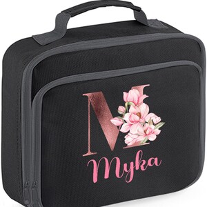 Puede incluir: Bolsa de almuerzo negra con asa y ribete grises. La parte delantera presenta la letra "M" en oro rosa con detalles florales rosas y el nombre "Myka" en escritura rosa. La bolsa tiene cierre de cremallera.