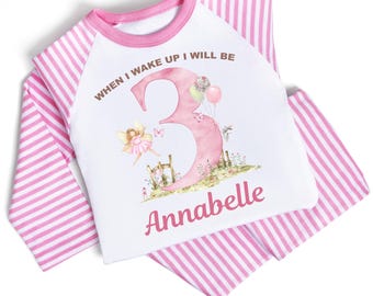 Puede incluir: Pijama a rayas rosa y blanco de manga larga. La parte superior tiene un fondo blanco con el texto "When I wake up I will be 3" y un número 3 rosa con un hada y mariposas. El nombre "Annabelle" está escrito debajo del número.