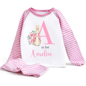 May include: Pink and white striped pyjamas with a white long-sleeved top featuring a pink letter A with a rabbit illustration and the text "is for Amelia".