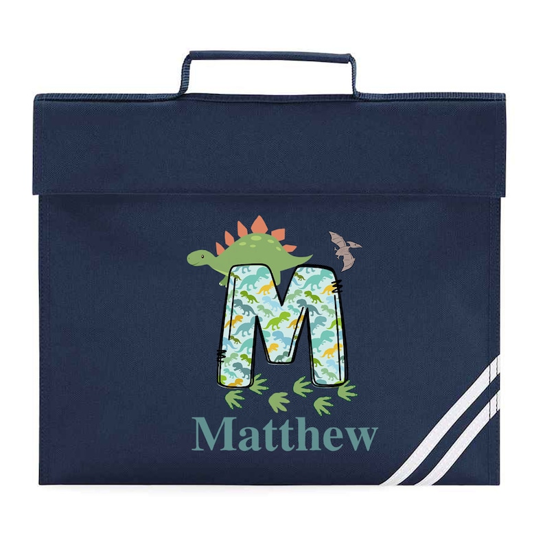 May include: A navy blue school bag with white stripes. The bag has a handle and features a dinosaur-themed design with the letter "M" and the name "Matthew" printed on it.