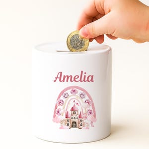 May include: White ceramic piggy bank with the name "Amelia" in pink script. A colorful rainbow and castle design decorates the front. A hand is placing a coin into the slot on top. The coin is a gold color.