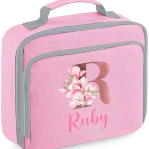 Puede incluir: Bolsa de almuerzo rosa con asa y ribete grises. La bolsa presenta una gran letra "R" en color oro rosa, con un dise&ntilde;o floral y el nombre "Ruby" impreso debajo. Tiene cierre de cremallera y bolsillo frontal.