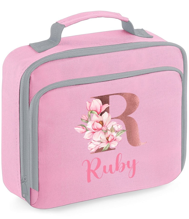Puede incluir: Bolsa de almuerzo rosa con asa y ribete grises. La bolsa presenta una gran letra "R" en color oro rosa, con un dise&ntilde;o floral y el nombre "Ruby" impreso debajo. Tiene cierre de cremallera y bolsillo frontal.