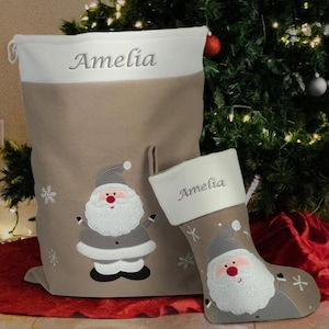 May include: A beige Christmas stocking and gift bag, both embroidered with the name "Amelia" in grey. They feature a Santa Claus design and snowflakes. The stocking and bag are on a red surface, with a Christmas tree in the background.