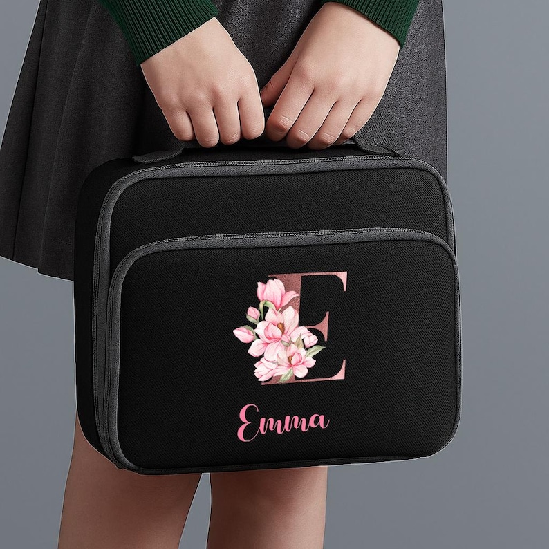 Puede incluir: Bolsa de almuerzo negra con ribete gris, con un dise&ntilde;o floral y la letra "E" en rosa. El nombre "Emma" est&aacute; escrito en rosa debajo del dise&ntilde;o floral. La bolsa tiene un asa en la parte superior.