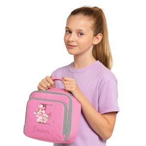 Puede incluir: Una bolsa de almuerzo rosa con ribete y asa grises. La bolsa presenta un dise&ntilde;o floral con la letra "E" y el nombre "Emma" en cursiva. La bolsa es sostenida por una persona que lleva una camiseta de color lila claro.