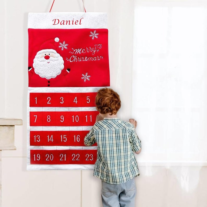 May include: A red and white Christmas advent calendar with the name "Daniel" at the top. The calendar features a Santa Claus design and the words "merry Christmas". Numbers 1-23 are displayed on the calendar. A child is looking at the calendar.