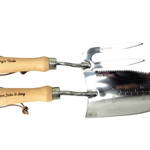 May include: A set of two gardening tools with wooden handles. The handles are engraved with the text "Nanny's Tools" and "Love From Jake & Amy". The tools are made of stainless steel and include a trowel with a measuring scale in inches and a hand rake.
