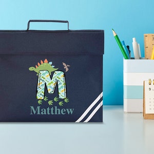 May include: A navy blue book bag with a white stripe design. The bag features a dinosaur and the letter M with a dinosaur pattern. The name Matthew is printed below the letter.