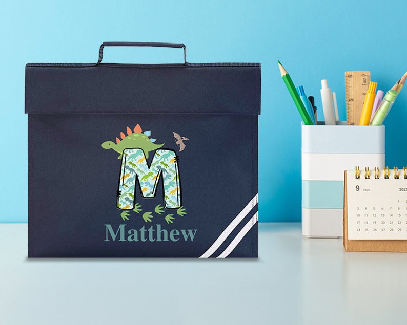 May include: A navy blue book bag with a white stripe design. The bag features a dinosaur and the letter M with a dinosaur pattern. The name Matthew is printed below the letter.
