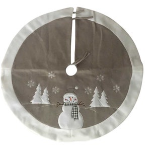 May include: A circular Christmas tree skirt in taupe with a wide white border. The centre features an embroidered white snowman wearing a black and white buffalo check scarf, flanked by white pine trees and scattered white snowflake patterns.