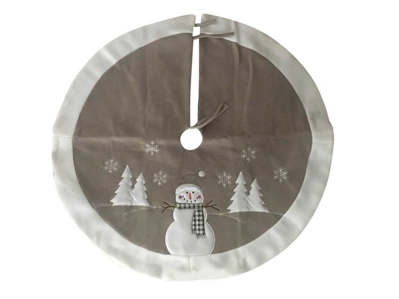 May include: A circular Christmas tree skirt in taupe with a wide white border. The centre features an embroidered white snowman wearing a black and white buffalo check scarf, flanked by white pine trees and scattered white snowflake patterns.