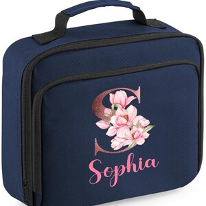 Puede incluir: Bolsa de almuerzo azul marino con asa y cremallera negras. La parte delantera presenta un dise&ntilde;o floral con la letra "S" y el nombre "Sophia" en rosa. Dise&ntilde;ada para transportar alimentos y bebidas.