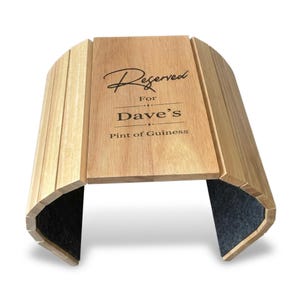 May include: Wooden beer caddy with the text "Reserved For Dave's Pint of Guinness" engraved on the front. The caddy is made of light-coloured wood with a curved design and a dark grey felt base.