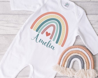 Puede incluir: Body blanco para bebé con un diseño de arcoíris colorido y el nombre "Amelia" en escritura turquesa. Un pequeño corazón rojo está encima del nombre. Un arcoíris de macramé decora la esquina inferior derecha.