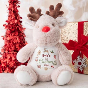 May include: A plush reindeer toy with brown antlers, a red nose, and a white belly. The belly features the text "Evie's 1st Christmas 2026" surrounded by Christmas-themed illustrations. A red Christmas tree and a wrapped gift are in the background.