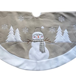 May include: A semi-circular Christmas tree skirt in a soft beige fabric with a wide white border. The design features an embroidered white snowman wearing a plaid scarf and hat, surrounded by white embroidered pine trees and snowflakes.