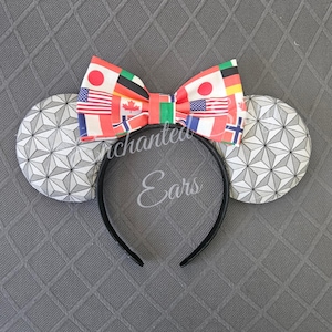 May include: Black headband with mouse ears. The ears are white with a gray geometric pattern. A bow with flags of various countries is centered on the headband. The text "Enchanted Ears" is printed on the headband.